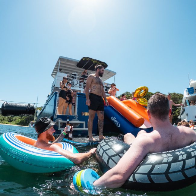 People enjoying a sunny day on a lake with a houseboat featuring a slide and canopy. Some are in the water on various inflatables, while others relax on the boat, making it perfect for Corporate boat events Sydney.