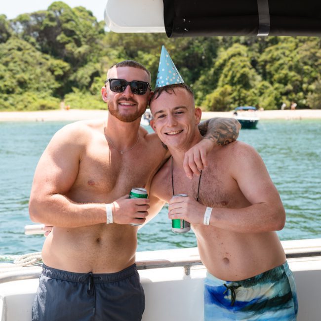 Two shirtless men on a boat, one wearing a birthday hat. Both are smiling and holding cans, with green foliage and water in the background. Enjoy moments like these with a private yacht charter in Sydney Harbour.