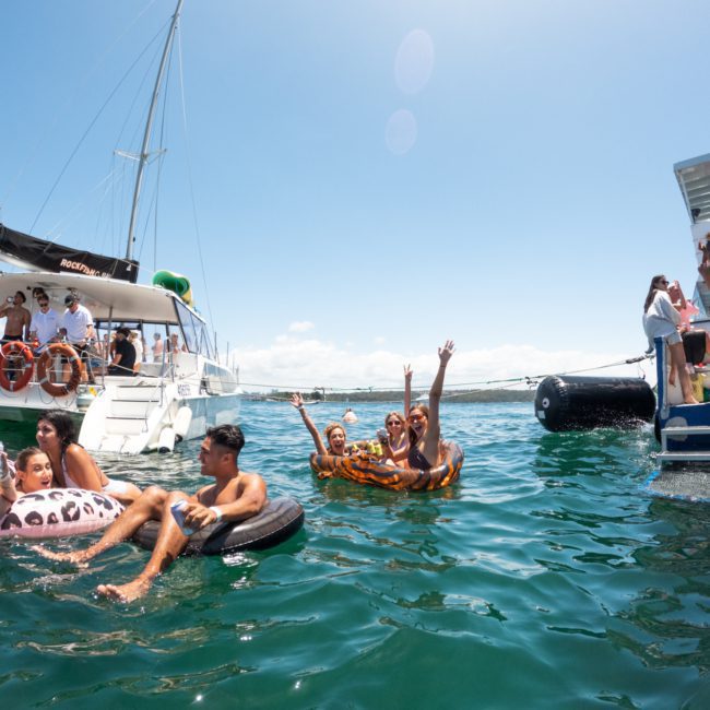 People lounging on inflatables and boats in the water, enjoying a sunny day. Some are waving, while others socialize and relax at a vibrant Sydney boat party hire.
