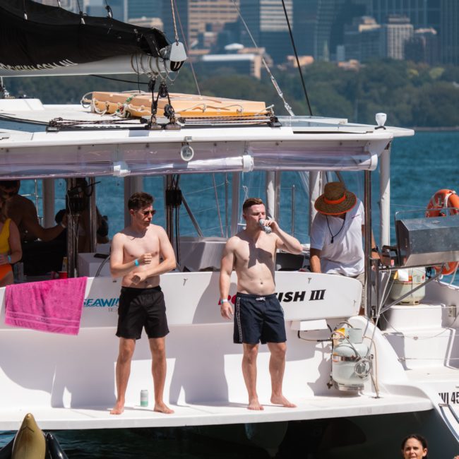 Several people enjoy a sunny day on a boat and in the water. Two shirtless men stand on the deck of this luxury yacht hire in Sydney, one drinking a beverage. Buildings are visible in the background.