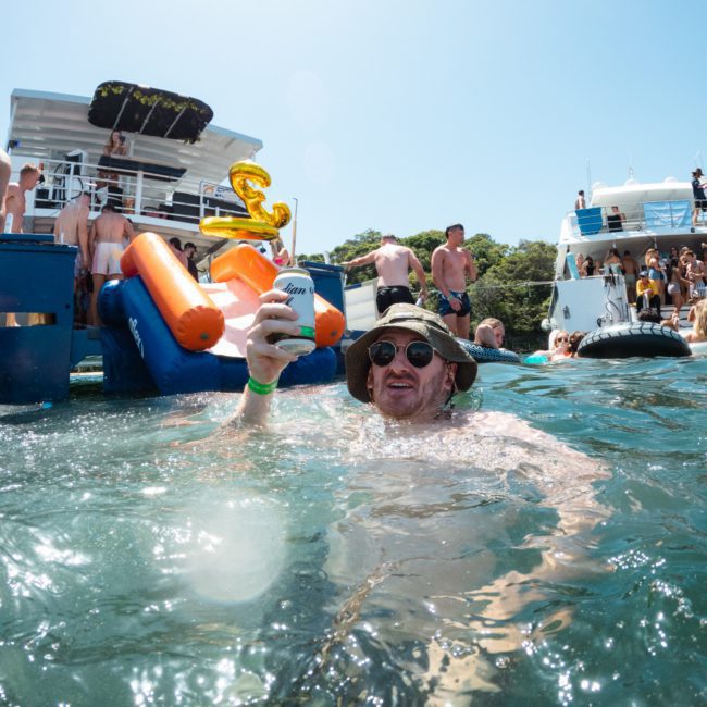 A person in a bucket hat and sunglasses holds a drink while standing in the water near boats with slides and people aboard on a sunny day, enjoying the vibes of a Catamaran party Sydney.