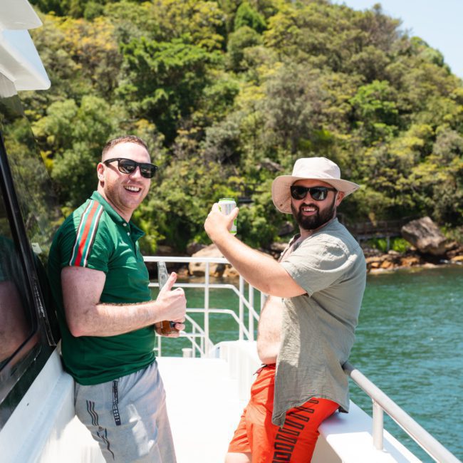 Two men, one in a green shirt and the other in a gray shirt and hat, stand on the deck of a boat near lush, green trees. The man in red shorts flexes his arm while the other holds a drink and smiles, enjoying their luxury yacht hire in Sydney Harbour.