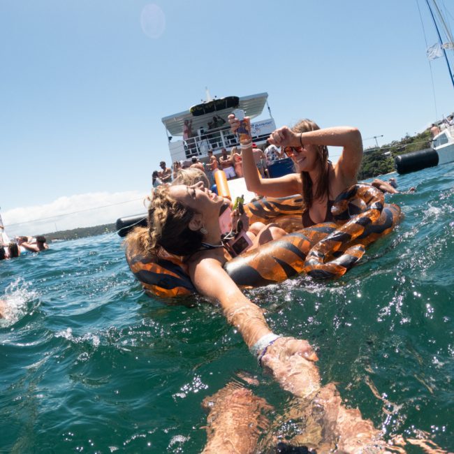 Two people relax on an inflatable float in the water, smiling and holding drinks during a Luxury yacht hire Sydney. In the background, a boat with several people adds to the festive atmosphere.