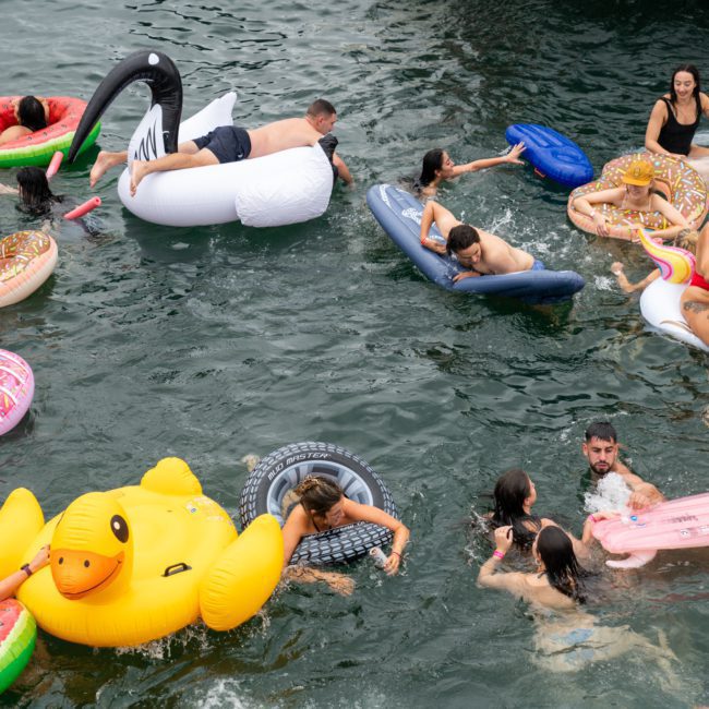 A group of people are swimming and floating on inflatable pool toys in a body of water, including a unicorn, swan, and duck, while nearby a luxury yacht hire Sydney hosts an exclusive event.