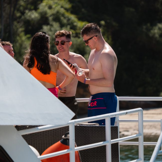 Three people in swimwear are standing on a boat, engaged in conversation. Trees and a beach appear in the background, creating an ideal setting for a private yacht charter on Sydney Harbour.