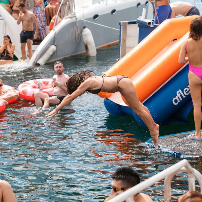 A person in a dark bikini is diving off a blue and orange slide into the water from a yacht, surrounded by other people swimming and lounging on inflatables at a lively Catamaran party in Sydney.