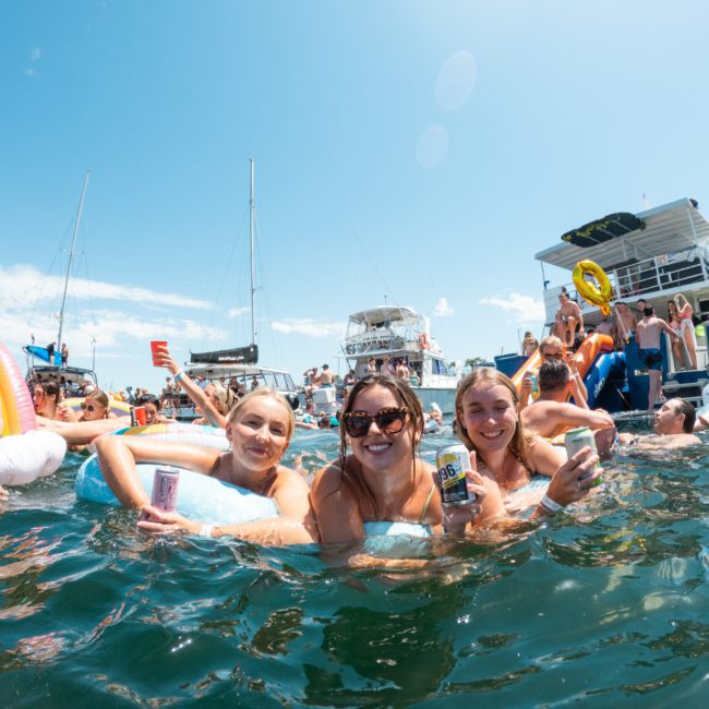 A group of people enjoying a sunny day in the water, holding drinks with a luxury yacht hire Sydney and other individuals in the background.