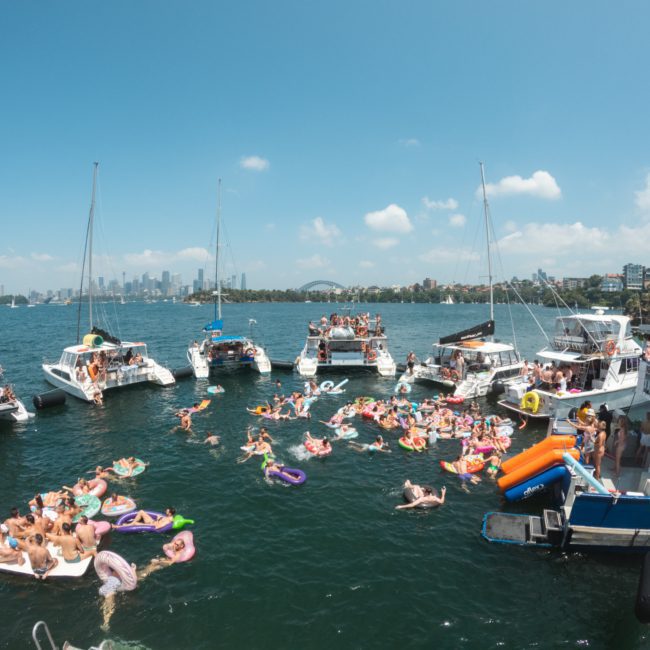 Boats are anchored close together in a bay, with people on colorful inflatables and floating devices enjoying a social gathering in the water. City skyline and trees are visible in the background, creating an idyllic setting for Sydney boat party hire.
