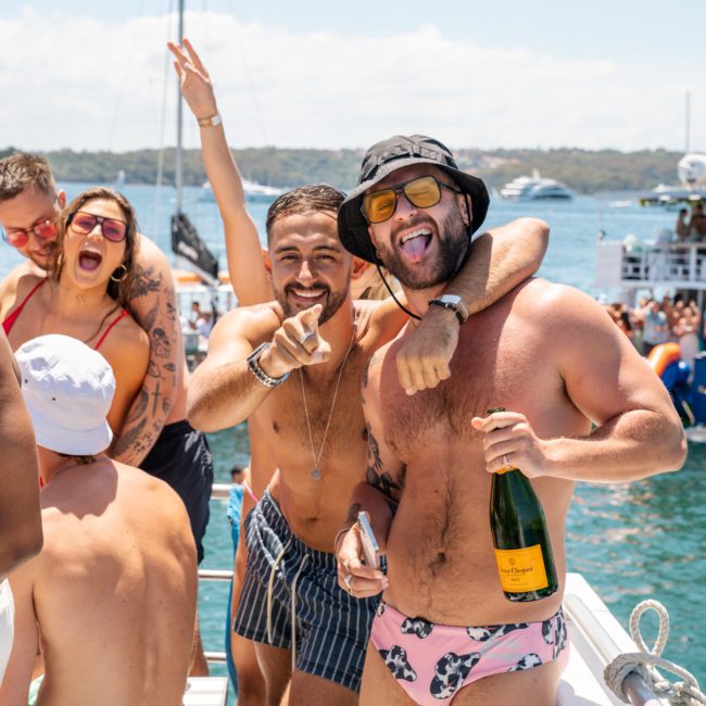 A group of people enjoying a sunny day on a boat, with two men in swimwear posing cheerfully, one holding a champagne bottle. The backdrop reveals sparkling water and smiling faces. Perfect for those considering Sydney boat party hire for their next event.