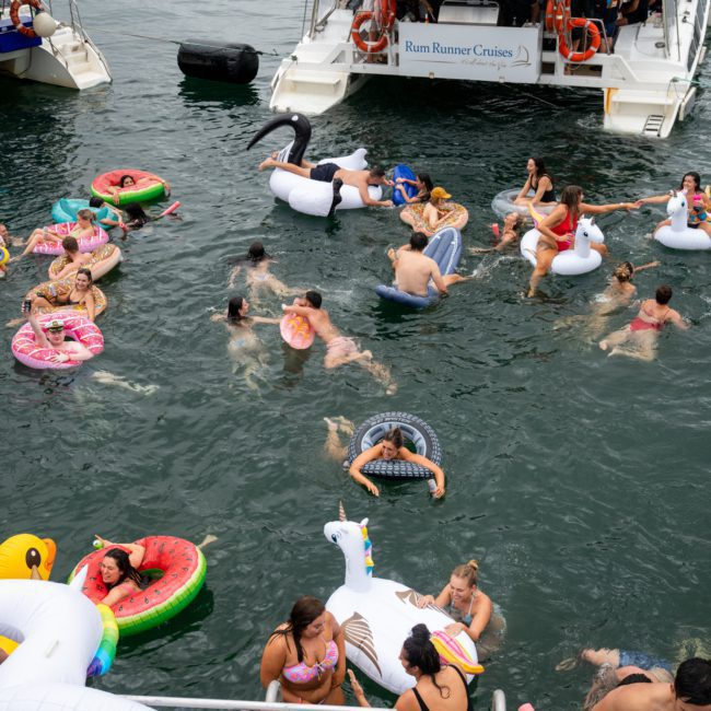 People swimming in a body of water with various inflatable pool toys, near two boats named "Rum Runner Cruises," enjoying a Sydney boat party hire.