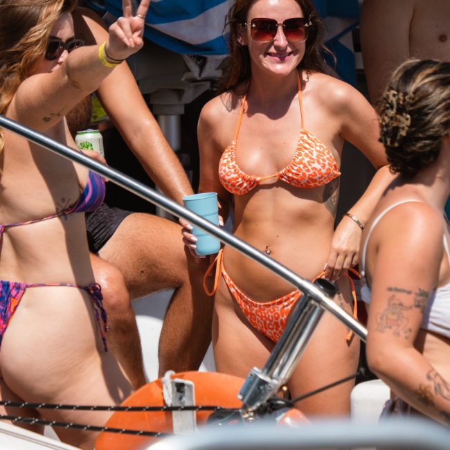 A group of people in swimwear enjoys a sunny day on a luxury yacht hire in Sydney. One person flashes a peace sign, and another holds a blue cup. A striped towel is seen in the background.