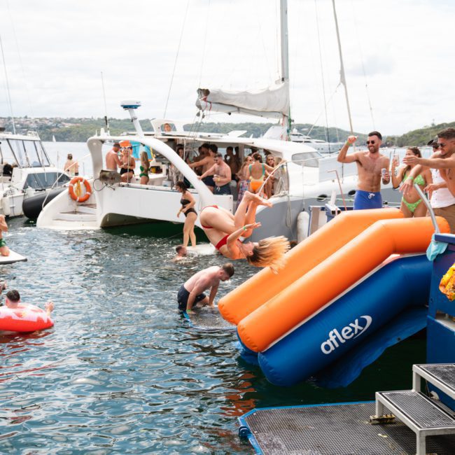 People enjoy water activities on a sunny day, with some on boats and others in the water. A woman is caught mid-flip off an orange and blue inflatable slide, while others cheer and watch. It’s perfect for a catamaran party in Sydney Harbour or even a private yacht charter adventure.