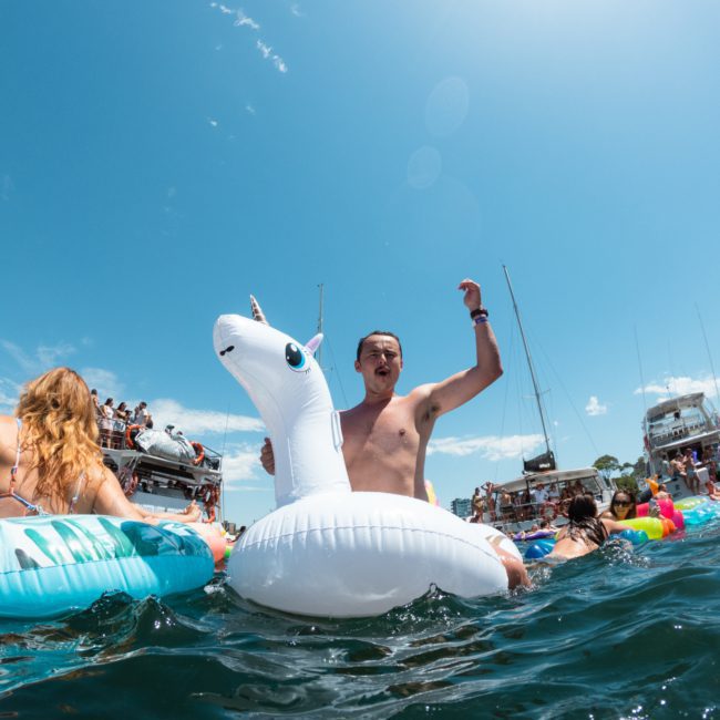 A man is floating on an inflatable unicorn in the water, raising one arm. Other people and boats are visible in the background under a clear blue sky, possibly enjoying a DJ boat hire Sydney nearby.