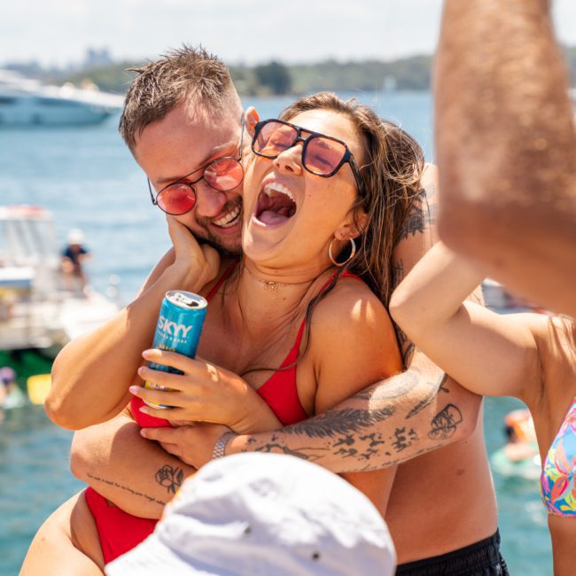 A man and woman laugh and embrace at an outdoor waterfront event. The woman holds a canned beverage while both wear sunglasses. Other people and boats, including a private yacht charter, are visible in the background.