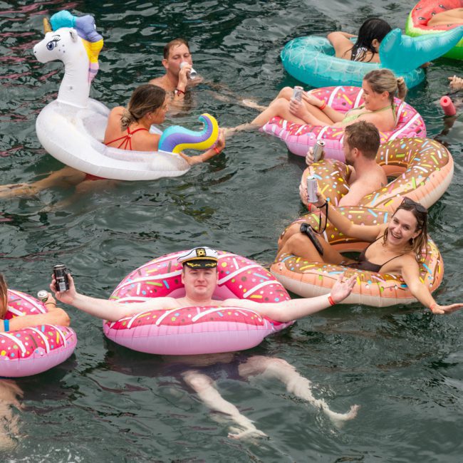 People floating in a body of water on inflatable pool floats, including a unicorn, donuts, and round floats, some holding drinks and socializing. Enhance the fun by considering a Sydney boat party hire or a private yacht charter in Sydney Harbour for an unforgettable experience.
