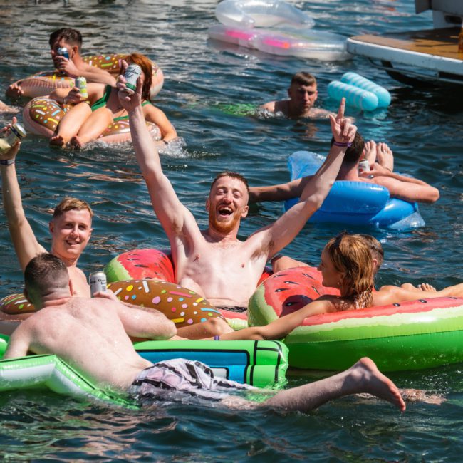 A group of people enjoying a sunny day in a body of water, floating on inflatable pool toys, including donut and watermelon designs. Some are holding beverages and smiling, while others are excited about their upcoming DJ boat hire Sydney experience.