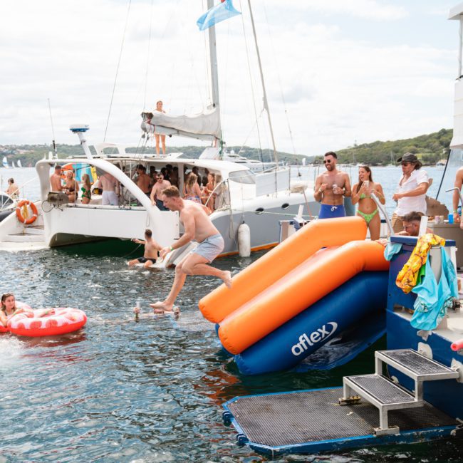 People are enjoying a luxury yacht hire in Sydney, with several anchored boats. Some individuals are sliding into the water from an inflatable slide, while others swim and float on inflatables.