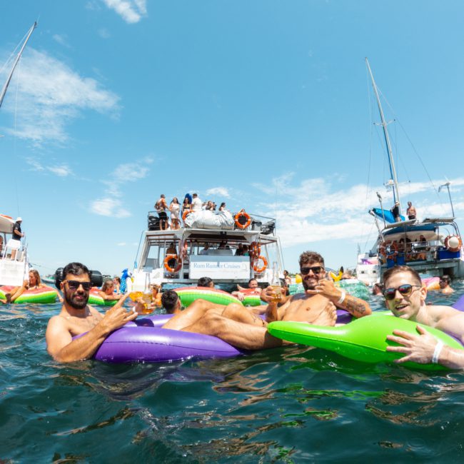 People relaxing on inflatable loungers in the water near boats on a sunny day. Some individuals are enjoying beverages while others are socializing on the boats in the background. It’s a perfect day for a Sydney boat party hire or even considering a private yacht charter Sydney Harbour to elevate your experience.