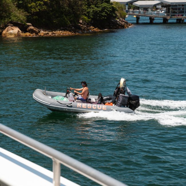 A small rescue boat with "RESCUE" written on the side speeds through the water, manned by two individuals wearing life vests. The shoreline and a dock are visible in the background, reminiscent of a Sydney boat party hire scene.