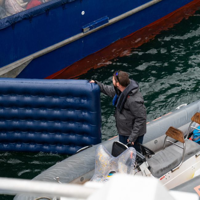 A man in a dark jacket stands in a small boat holding the edge of a blue inflatable mattress, positioned next to a larger vessel during an exclusive private yacht charter on Sydney Harbour.