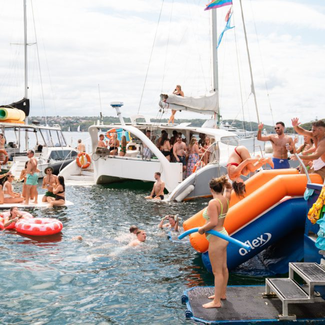 A group of people enjoy a day on the water, swimming and lounging on inflatables near several anchored boats. Some are on a blue and orange inflatable slide leading from a boat into the water, reveling in what's clearly an unforgettable catamaran party in Sydney.