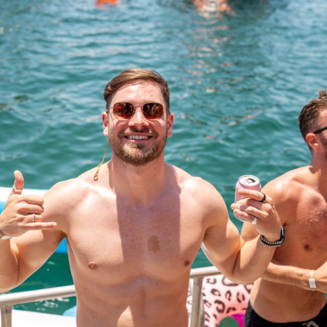 A shirtless man wearing sunglasses smiles and makes a hand gesture while holding a drink, standing on a boat at a Sydney boat party hire with water and other people in the background.