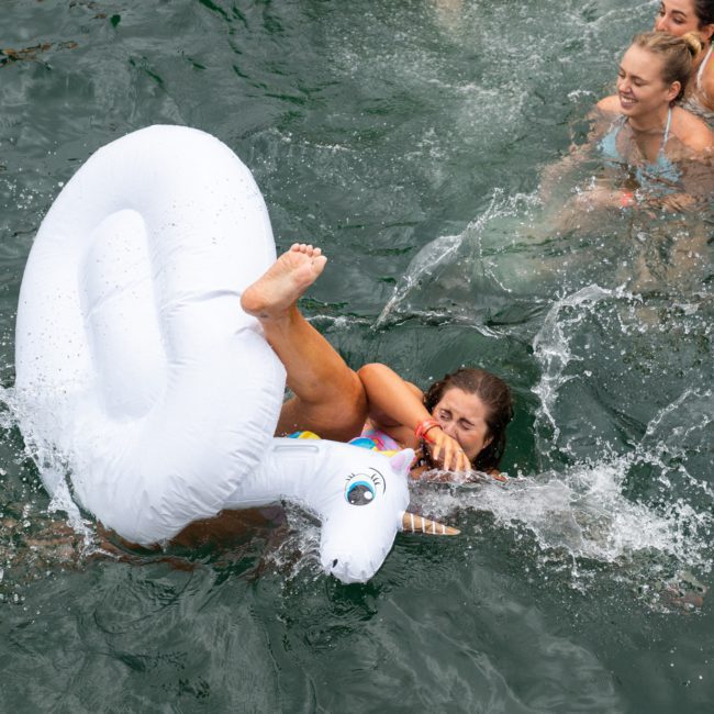 Three people are swimming in the water, one of them is trying to stay balanced on a white inflatable unicorn float, enjoying the fun vibes of a Sydney boat party hire.