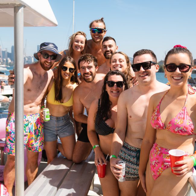 A group of people in swimwear are gathered together on a sunny day, posing for a photo on a private yacht charter in Sydney Harbour. They are smiling and holding drinks, enjoying the cruise.