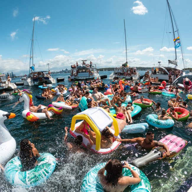 People on inflatable pool floats gather in water near anchored boats under a sunny sky, participating in a lively Sydney boat party hire.