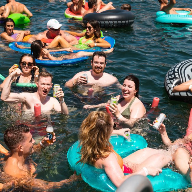 People floating on inflatable tubes and pool noodles in a body of water, enjoying beverages on a sunny day, near a DJ boat hire in Sydney.