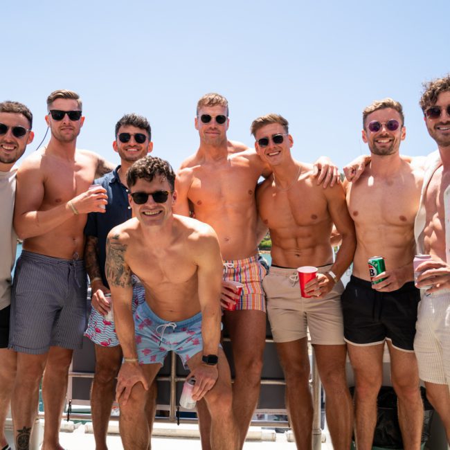 A group of eight men pose for a photo on a boat, most of them shirtless and wearing sunglasses. The background shows clear blue water and a sunny sky, perfectly capturing the vibe of a Sydney boat party hire.