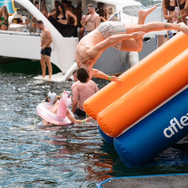 A group of people enjoy a water activity on a boat, with some swimming and others using an inflatable slide to enter the water. One person mid-air jumps from the slide during their private yacht charter Sydney Harbour.