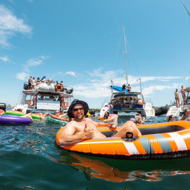People enjoying a sunny day on the water, floating on inflatable rafts and swimming near sailboats. A man in an orange and black raft gives a thumbs up while others socialize around, capturing the essence of a Sydney boat party hire.