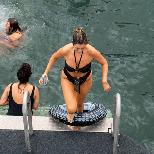 A woman in a black swimsuit climbs out of the water using a ladder, a tire-shaped float around her waist. Two other people are seen swimming nearby, enjoying the pristine waters at a private yacht charter Sydney Harbour event.
