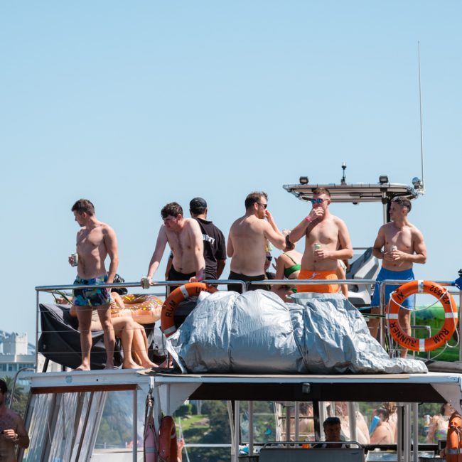 A group of people in swimwear stands on the deck of a luxury yacht hire Sydney, with life rings, enjoying a sunny day. A bridge is visible in the background.