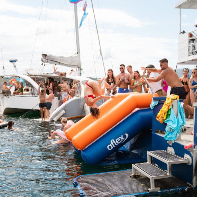 People are gathered on boats and in the water, with a person sliding down an inflatable slide from one boat. Others observe from the boats while some swim and paddleboard in the water, enjoying what appears to be a lively catamaran party in Sydney.