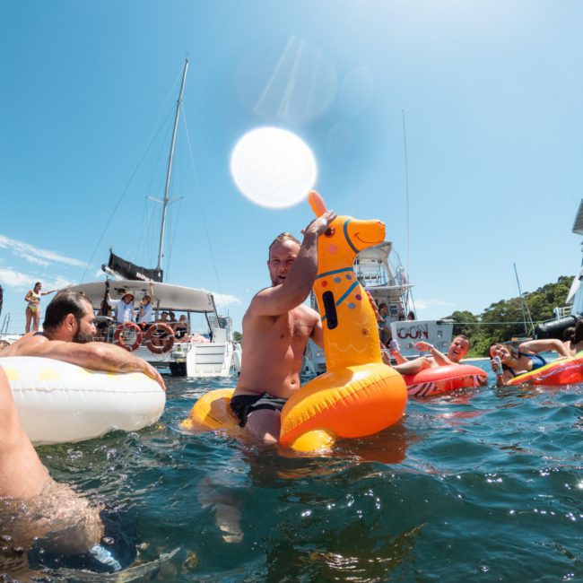 A man in swim trunks rides an inflatable giraffe float in the water, surrounded by other people on floatation devices with boats from a Sydney boat party hire in the background under a clear blue sky.
