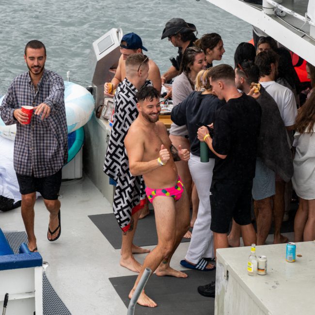 A group of people enjoying a Catamaran party Sydney on a boat, with one man in a colorful swimsuit dancing energetically in the center.