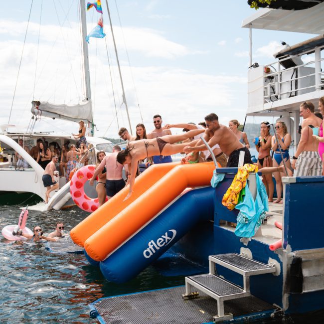 People in swimsuits having fun on a boat, with some descending orange water slides into a lake. Two boats are docked, and inflatable floats are visible in the water during an exhilarating Sydney boat party hire.
