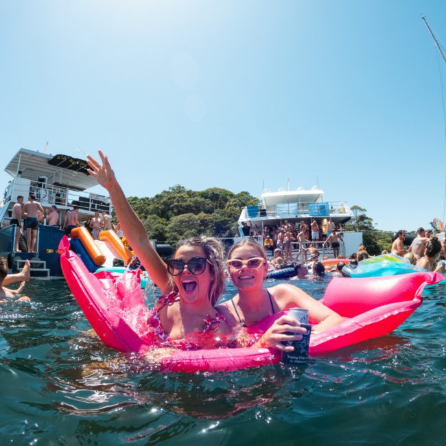 Two people enjoying a sunny day on a pink float in the water, surrounded by boats and others swimming and relaxing. Nearby, a luxury yacht hire in Sydney Harbour adds an extra touch of elegance to the scene.