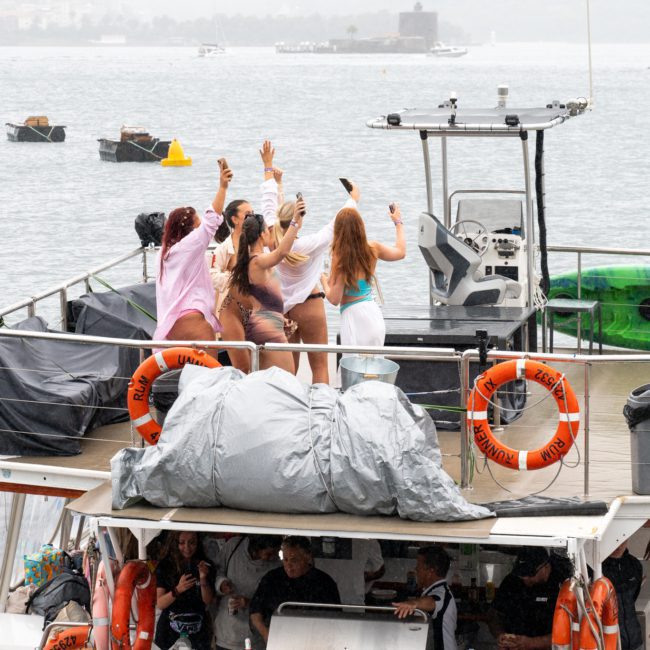 A group of people on the deck of a boat are waving and dancing near the water, with life rings, tarps, and a kayak visible – all part of an exciting corporate boat event in Sydney.