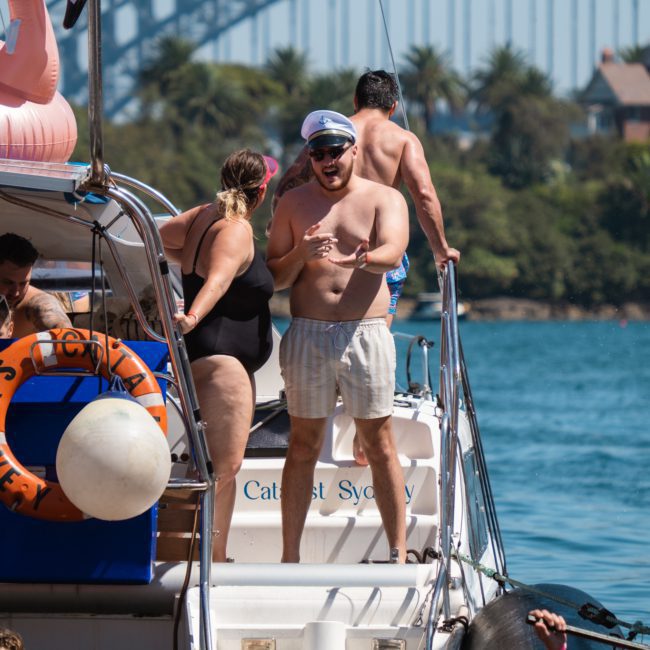 People on a luxury yacht with a flamingo float in the water, near a bridge. One person wearing a captain's hat and swimming trunks gestures with his hand, while others sit or stand on the yacht. This private yacht charter is perfect for enjoying Sydney Harbour’s beautiful scenery.