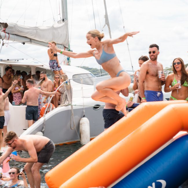 A group of people enjoy a Sydney boat party hire. A woman in a blue swimsuit jumps from an inflatable slide into the water, while others watch and socialize nearby.