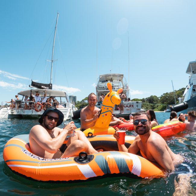 Three men are enjoying drinks while floating on inflatable rafts in a body of water, with boats and other people in the background on a sunny day. It captures the essence of a catamaran party in Sydney, creating an atmosphere perfect for leisure.
