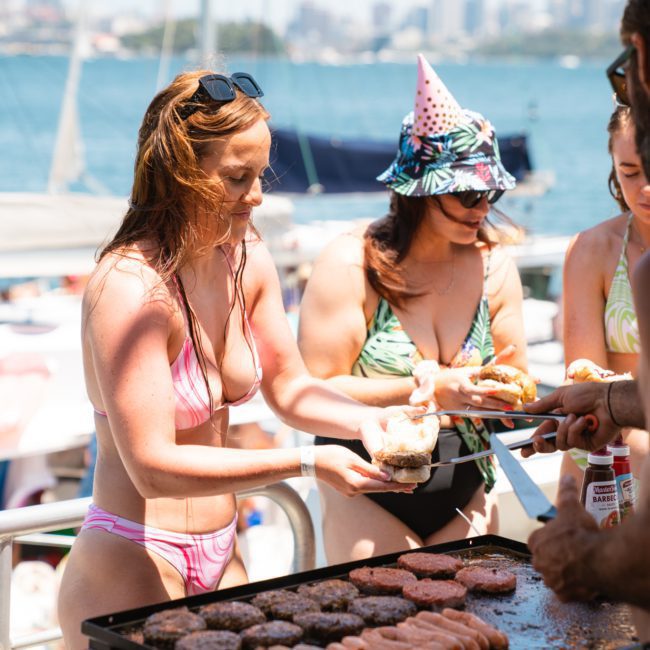People serving themselves food from a grill on a luxury yacht hire Sydney, with a scenic waterfront background.