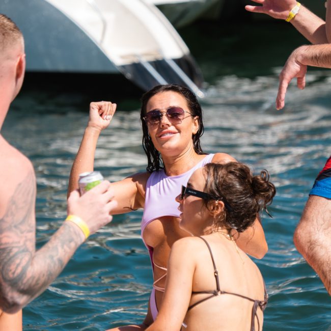 A group of people enjoying a sunny day on a boat, with one woman in front wearing a pale purple swimsuit and sunglasses, others around her holding drinks. This scene captures the essence of Sydney boat party hire, perfect for anyone looking to make the most of their time on the water.