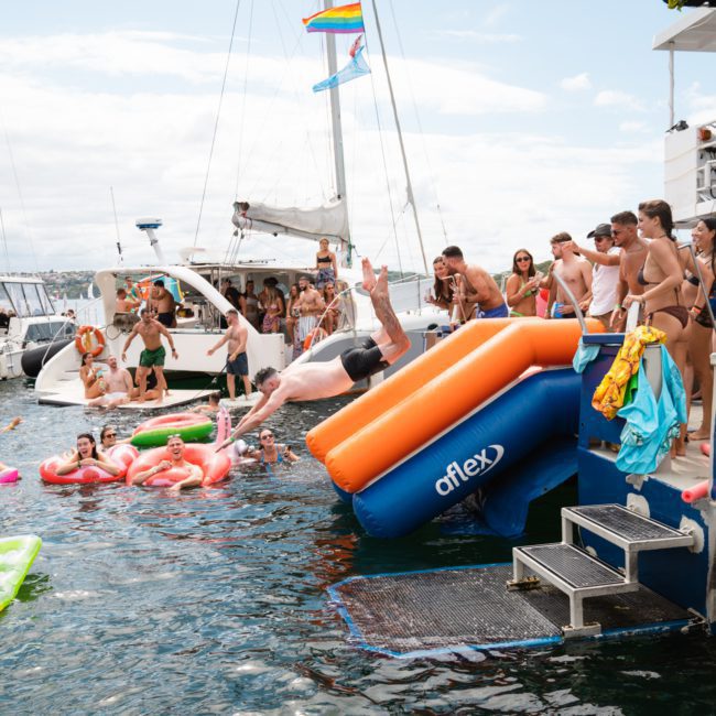 People on boats and in the water enjoy a sunny day. One person dives off an inflatable slide on a docked boat, while others swim and relax on inflatables, part of a lively Sydney boat party hire experience.