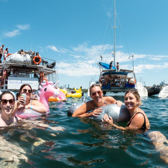 A group of people swim in the water near anchored boats on a sunny day, enjoying a lively Sydney boat party hire experience. Some hold inflatable toys, including a unicorn float. Several boats and other swimmers are visible in the background.