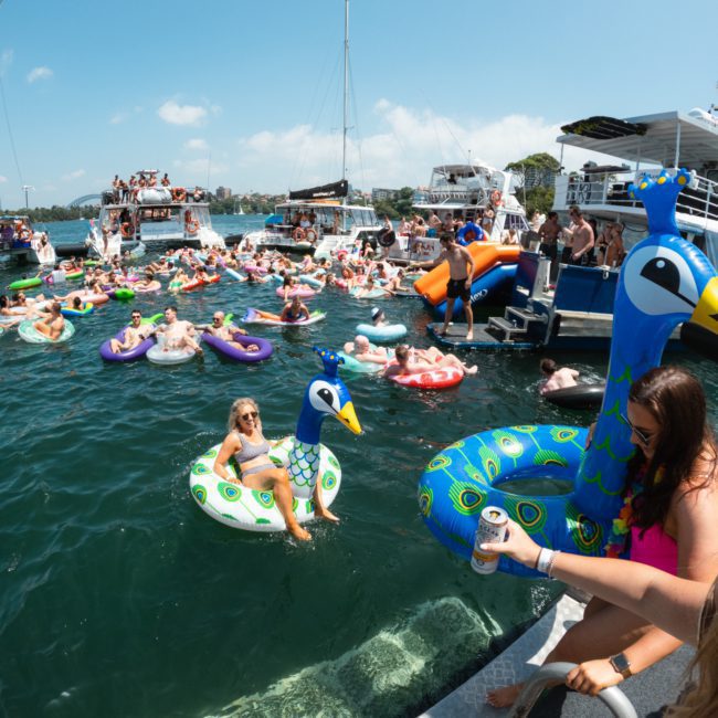 People are enjoying a sunny day on a lake, floating on inflatable pool toys like peacocks, with boats docked nearby. Some individuals are sitting by the dock, holding drinks and chatting about the upcoming catamaran party in Sydney.