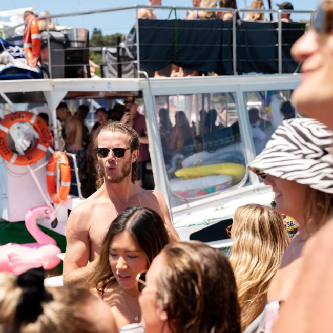 A group of people in swimsuits are on or near a boat during a sunny day. Some are standing, talking, and holding drinks. The background shows more people on the boat and various flotation devices, evoking the ambiance of a Luxury yacht hire Sydney experience.